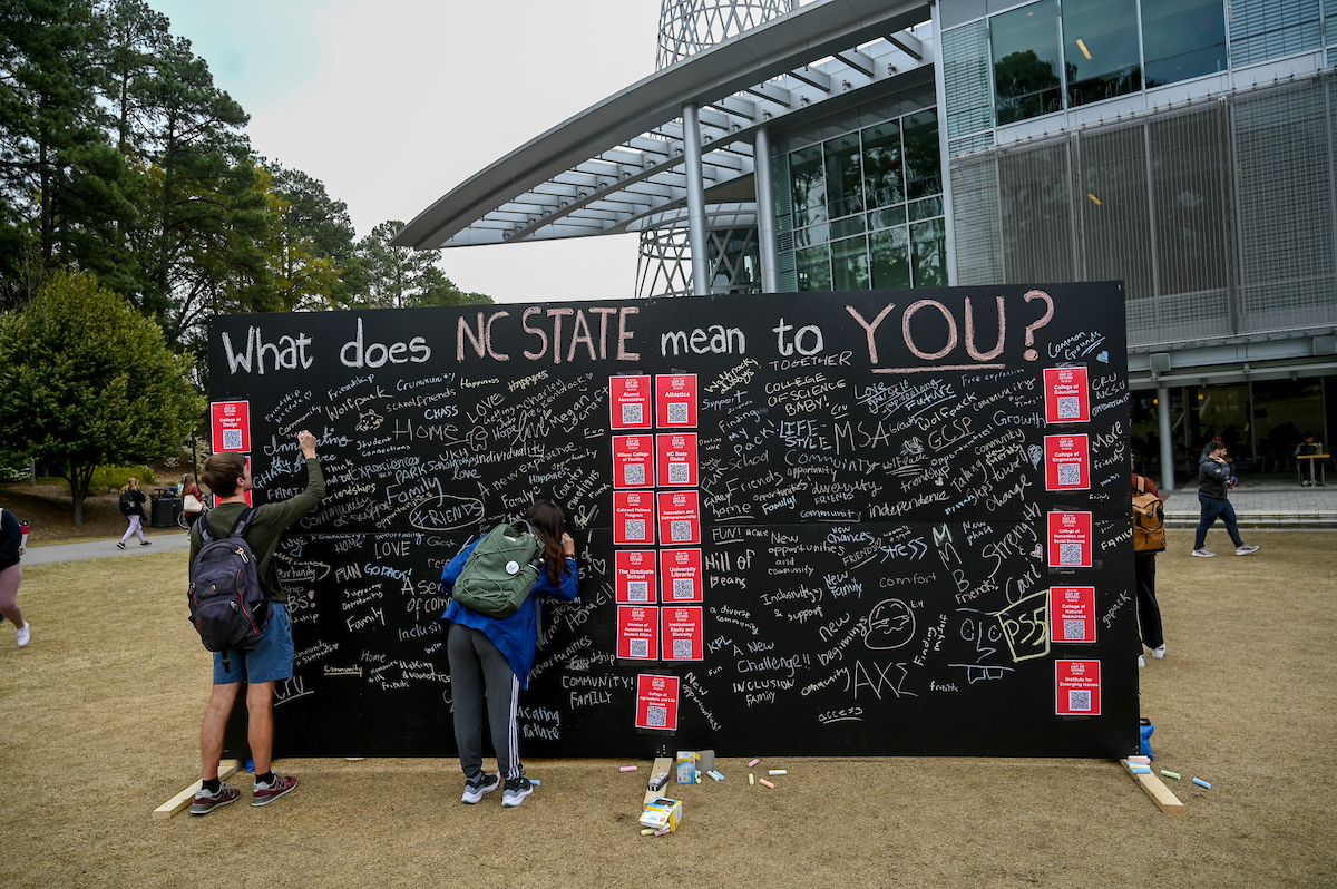 During Day of Giving, two students write on a large chalkboard with the title "What does NC State mean to you?"outside Talley Student Union.