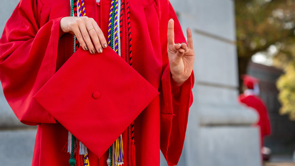 A student wearing red robes holds up their graduation cap in front of the Memorial Belltower.